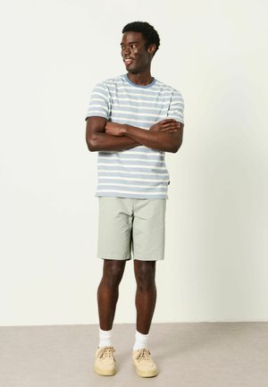 Mann steht mit verschränkten Armen, trägt ein hellblau-weiß gestreiftes T-Shirt, beige Shorts, weiße Socken und sandfarbene Turnschuhe.