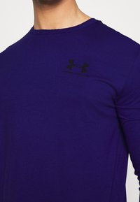 Man som bär en mörkblå Under Armour långärmad tröja med logotypen på den övre vänstra sidan av bröstet, framför en enfärgad bakgrund.