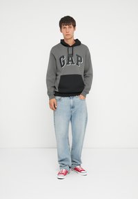 Hanorac gri și negru cu logo-ul "GAP", combinat cu blugi albaștri deschiși și adidași roșii. Stil casual, croială lejeră, cu glugă ajustabilă.