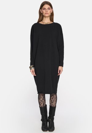 Kobieta ubrana w luźną czarną sukienkę z długim rękawem, wzorzyste czarne rajstopy, czarne kozaki do połowy łydki, bransoletki i złoty naszyjnik, stojąca na białym tle.