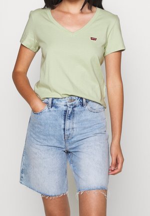 Kvinde iført lysegrøn V-hals Levi's T-shirt og lyseblå højttaljede denimshorts med frynsede kanter, den ene hånd i lommen.