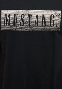 Tessuto nero con una toppa rettangolare bianca che mostra la parola "MUSTANG" in caratteri alti, neri e consumati.