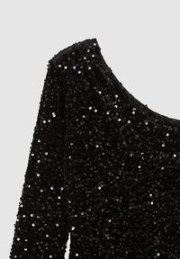 Dettaglio di tessuto ricoperto di paillettes nere che mostra una manica lunga e la zona della spalla di un indumento su uno sfondo chiaro e uniforme.
