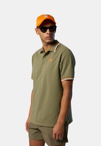 Polo shirt verde oliva con vestibilità classica, caratterizzato da un colletto solido con strisce arancioni, bianche e nere. Abbinato a un berretto arancione e occhiali da sole.