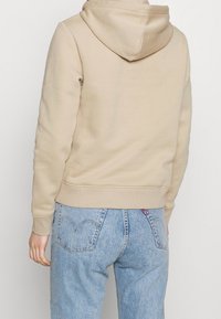 Felpa beige in cotone con cappuccio, polsini e orlo a costine. Indossata con jeans in denim azzurro chiaro con dettagli sulle tasche posteriori.