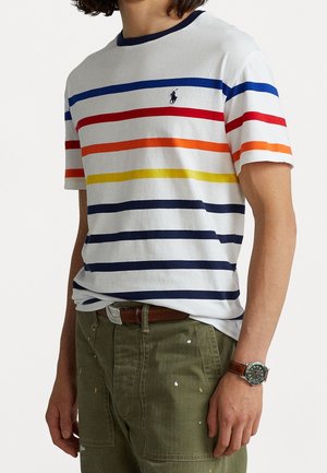 Man wearing white T-shirt with horizontal blue, red, orange, and yellow stripes, olive green pants, and brown leather watch.
