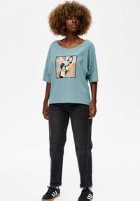 Lichtblauw oversized t-shirt met een vierkante graphic van een hond, gecombineerd met donkere jeans en zwarte sneakers, met witte en tan accenten.
