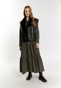 Zwarte leren vest met een faux fur kraag, gelegd over een donkere bloemenprint jurk. Model draagt zwarte enkellaarsjes. Soepele textuur en gestructureerde vorm.