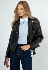 Giubbotto in pelle nera da motociclista con accenti di borchie argentate, tasche con zip e una cintura. Abbinato a una camicia colletto azzurro.