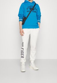 Felpa blu con lettering "HILFIGER NEW YORK", abbinata a pantaloni jogging bianchi con stampa "HIL FIGER". Comprende anche sneakers bianche.