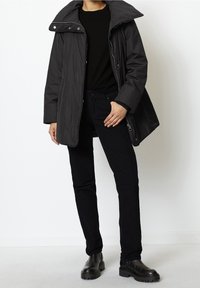Černý prošívaný kabát s vysokým límcem, dlouhými rukávy a zapínáním na zip, doplněný černými manšestrovými džínami a robustními černými botami.