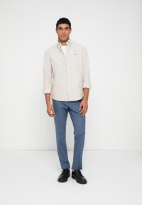 Men's light beige linen shirt with long sleeves, paired with blue trousers and black shoes. Simple, button-up design with a crisp look.