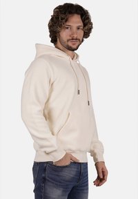 Felpa in pile color crema con tasca frontale, cappuccio con cordino e polsini a coste. Indossata con jeans blu, che evidenziano un look casual e una piacevole texture.