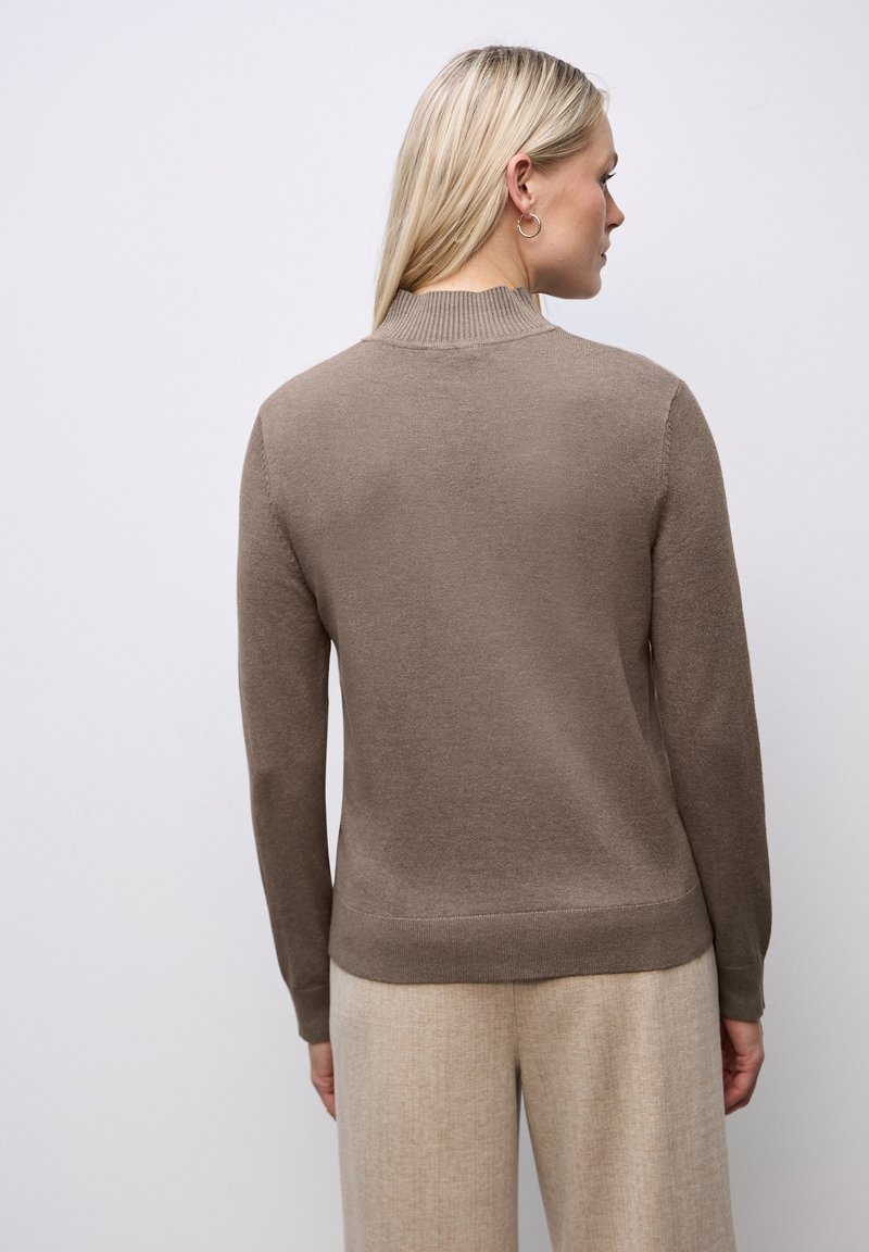 Kermanneule tan väri, jossa on korkea ribbo collar, pitkät hihat ja kapeat helmat. Pehmeä rakenne, sileä pinta ja minimalistinen muotoilu.