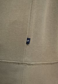 Tessuto verde oliva con una texture a coste, caratterizzato da un piccolo etichetta blu navy con un logo bianco. I dettagli delle cuciture sono visibili lungo i bordi.