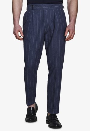 Dopasowane spodnie w granatowy prążek z bocznymi regulatorami noszone przez stojącą osobę w czarnych skórzanych mokasynach.