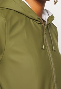 Veste à capuche vert olive avec une texture lisse et brillante, dotée d'une fermeture éclair, d'un cordon de serrage et d'une doublure intérieure blanche.