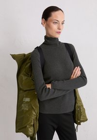 Grijze geribbelde coltrui gecombineerd met een gewatteerde olijfgroene jas die over de schouder hangt, met zwarte schouderbanden.