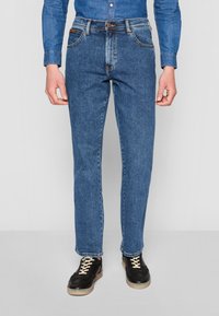 Jean en denim bleu moyen avec une coupe droite, comportant cinq poches, une braguette à boutons et des coutures contrastées. Porté avec des baskets noires.