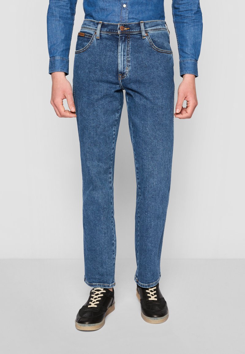 Jean en denim bleu moyen avec une coupe droite, comportant cinq poches, une braguette à boutons et des coutures contrastées. Porté avec des baskets noires.