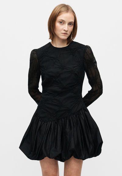 Rochie neagră cu un corpiță texturată, împodobită cu broderii, mâneci lungi transparente și o fustă evazată cu volane.