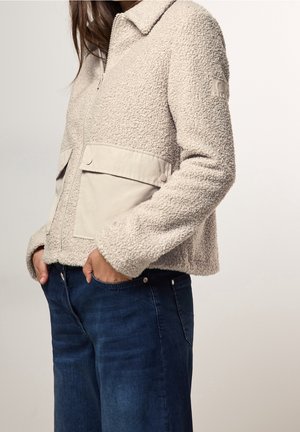 Frau trägt eine beige, strukturierte Jacke mit Reißverschluss und großen Vordertaschen sowie dunkelblaue Jeans, Hände in den Taschen, steht vor einem hellen Hintergrund.