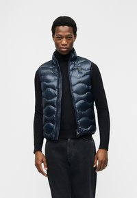 Άνδρας που φοράει μαύρο ζιβάγκο, μπλε σκούρο μπουφάν με πάλλευκα ραφές και μαύρο τζιν, στέκεται μπροστά σε λευκό απλό φόντο.