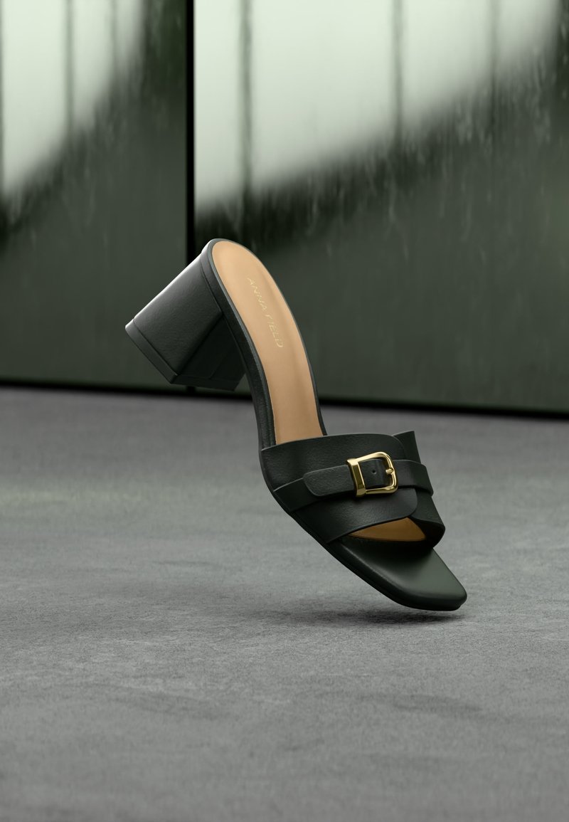 Mule en cuir noir à bout ouvert avec talon bloc et détail de boucle dorée, présenté sur une surface grise avec un fond sombre flou.