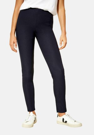 Femeie purtând pantaloni bleumarin mulați și adidași albi cu accente negre, stând cu brațele relaxate pe lângă corp.