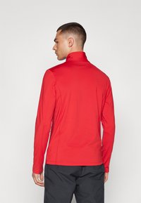 Haut de sport rouge à manches longues avec un col montant, fabriqué en tissu lisse ; présente un design ajusté et des détails de couture minimalistes.
