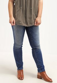 Henkilö, jolla on päällään haalistuneet siniset kapeat farkut, oliivinvihreä koristeltu paita ja ruskeat avonaiset korkokengät, seisoo valkoista taustaa vasten.