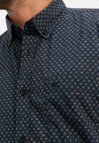 Blauw met patroon shirt met een button-down kraag. Het patroon bevat kleine oranje, blauwe en grijze vormen, met zwarte knopen en een subtiel logo.