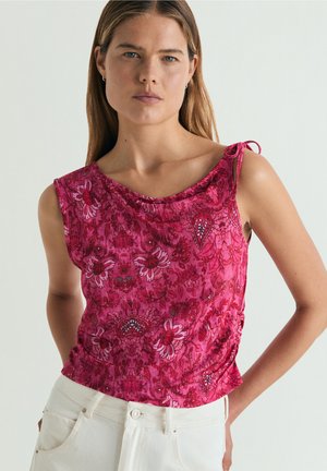 Femme portant un haut rose à fleurs sans manches avec un détail noué aux épaules et un pantalon clair, debout devant un fond uni.