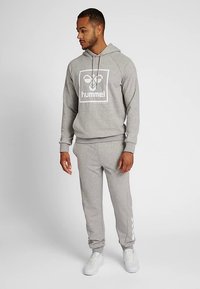 Grauer Kapuzenpullover mit einem zentralen weißen Logo, kombiniert mit passenden grauen Jogginghosen, die entlang des Beins ein weißes Streifendekor aufweisen.