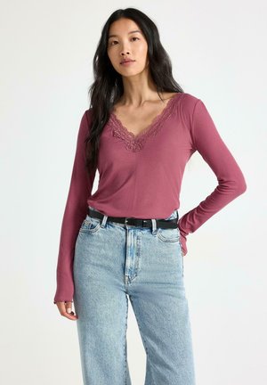 Kvinde med langt mørkt hår har en malvefarvet langærmet bluse med blondestrimler, der er stoppet ned i lyseblå højtaljede jeans med et sort bælte.