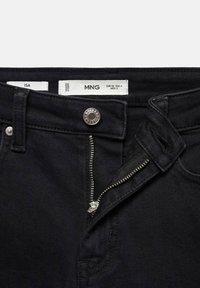 Jeans de mezclilla negros con cierre de botón plateado, cremallera y etiquetas de talla y marca. La vista en detalle enfatiza la textura de la tela.