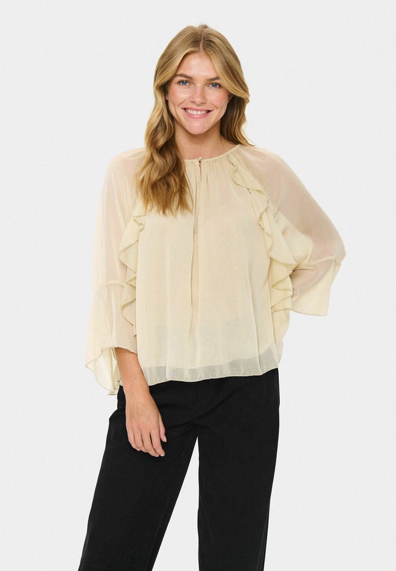 Blond kvinna som ler och bär en genomskinlig beige blus med volanger och svarta byxor, stående med ena handen på höften mot en vit bakgrund.