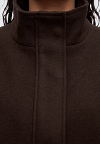 Gros plan d'un manteau marron foncé à col montant avec fermeture avant dissimulée et tissu doux et texturé visible autour du cou et des épaules.