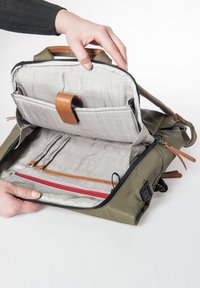Olivgrüner Rucksack mit einem Reißverschluss. Das Innere weist einen hellen Karostoff, Lederrandakzente und mehrere Organisationsfächer auf, darunter ein rotes Reißverschlussfach.