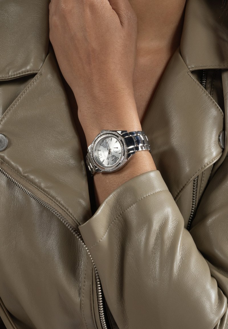 Poignet portant une montre en métal argenté avec un cadran rond et une lunette en cristal, reposant sur une main croisée sur la poitrine dans une veste en cuir taupe.