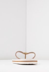 Bruine slippers met een hartvormige band, beige rubberen zool en een witte tussenzool met een bruine streep. Gouden accent op de band.