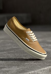 Beżowe sneakersy z płótna z białymi sznurówkami, czarną gumową podeszwą i kontrastującymi szwami. Posiadają naszywkę z logo z boku oraz teksturowaną górę.