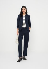 Ναυτικό μπλέ σακάκι με δύο πλαϊνές τσέπες, συνδυασμένο με ριγέ τοπ και ταιριαστό παντελόνι σε ναυτικό μπλε. Μαύρες μπότες με τακούνι από γυαλιστερό υλικό και λευκή τσάντα.