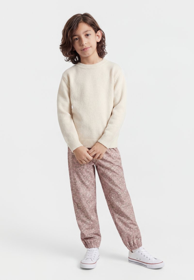 Niño con cabello castaño hasta los hombros, vestido con un suéter crema, pantalones jogger rosa con estampado y zapatillas blancas, de pie con las manos entrelazadas.