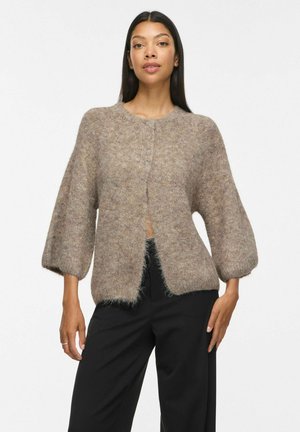 Femme portant un pull taupe duveteux boutonné avec manches ballon et un pantalon noir, debout, les mains reposant de chaque côté.