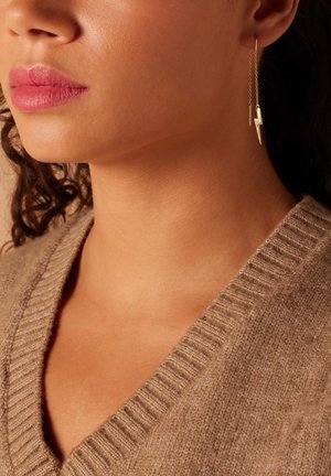 Boucles d'oreilles en plaqué or en forme d'éclair avec un design pendulaire, accompagnées d'un pull beige texturé avec un décolleté en V profond.
