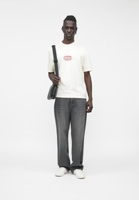 Camiseta de manga corta color beige claro con logo rojo, combinada con vaqueros de pierna ancha en gris oscuro y zapatillas blancas. Bolso negro de hombro colgado del brazo.