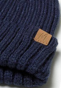 Strickmütze in Marineblau mit gerippter Textur, versehen mit einem quadratischen Lederpatch mit geprägtem "M". Weiche Wollmischung, faltbares Design.