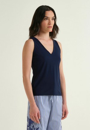 Femme aux cheveux foncés portant un haut sans manches bleu marine en V et un pantalon rayé bleu et blanc à imprimé floral, debout devant un fond uni.