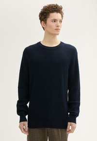 Granatowy sweter dzianinowy z teksturowanym wzorem w siatkę, okrągłym dekoltem i długimi rękawami. Manżety i dół wykończone są ściągaczem.